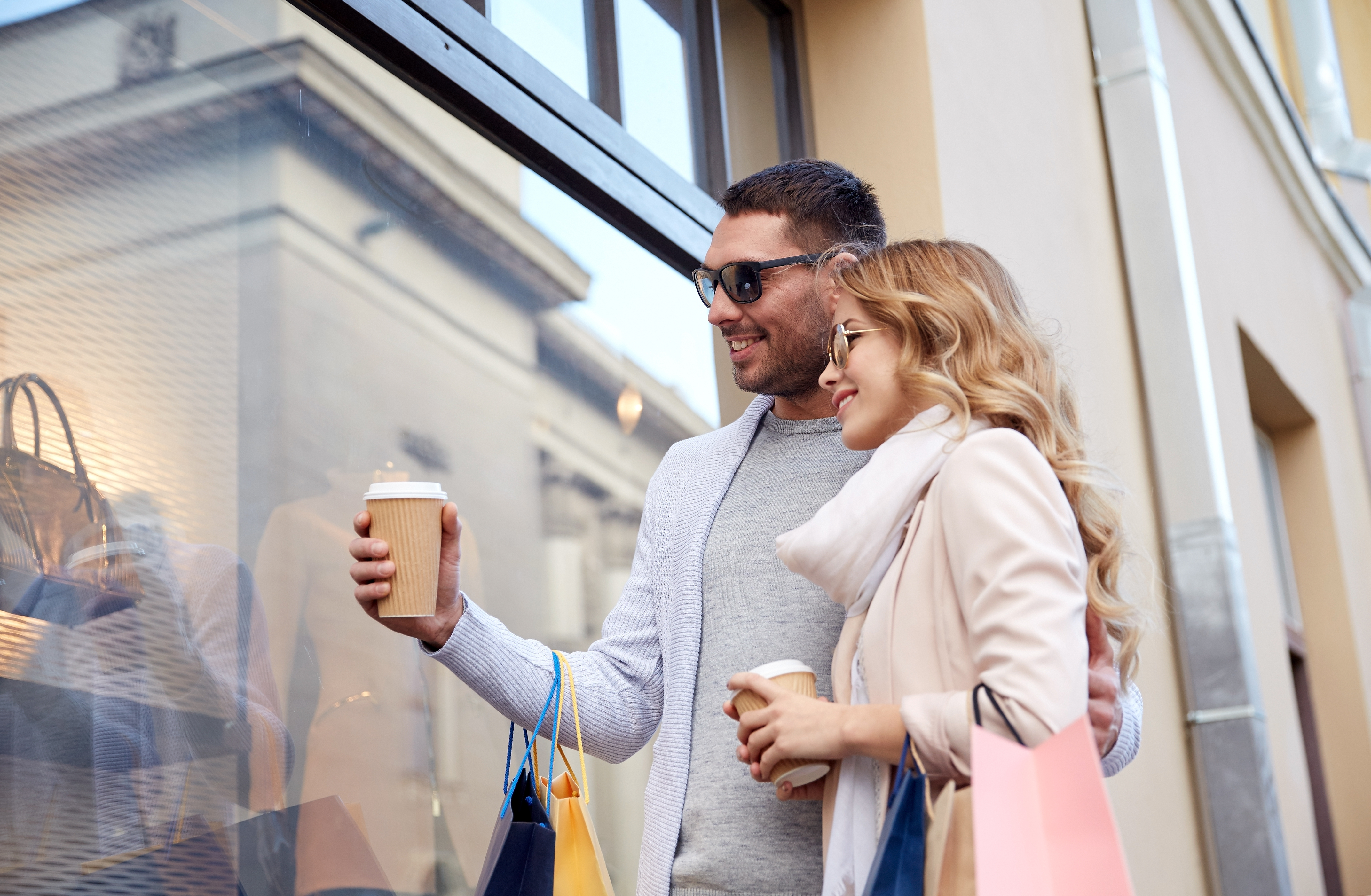 sale, consumerism and people concept - happy couple with shopping bags and coffee paper cups looking at shop window in city