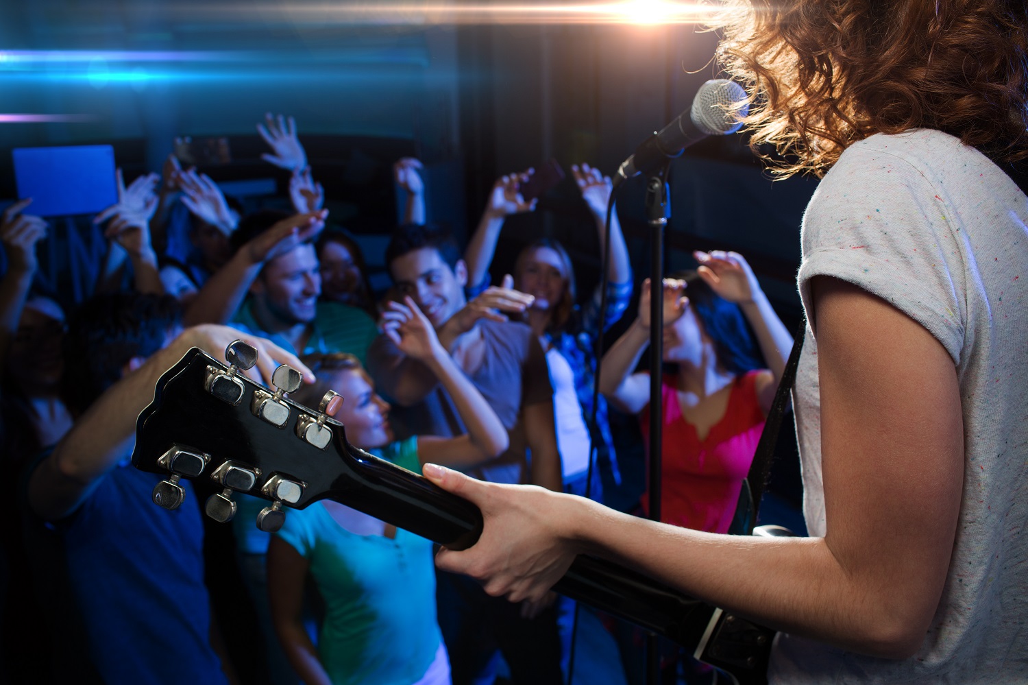 Close up of singer playing electric guitar and singing on stage over happy fans crowd waving hands at concert in night club