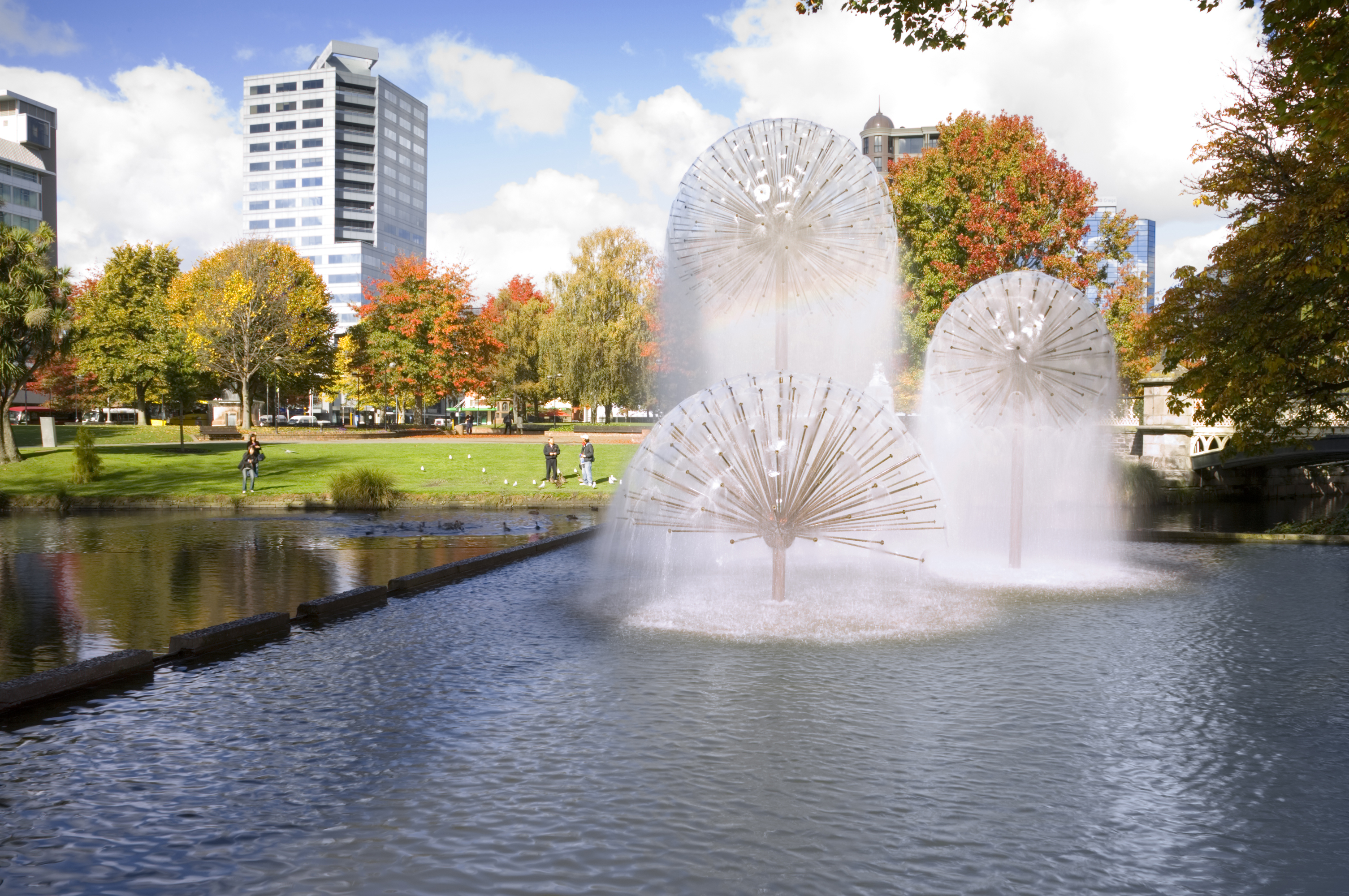 A sunny autumn day in Christchurch, New Zealand, with the Ferrier Fountain, the Avon River, tourists chatting and feeding ducks in background, and trees in autumn colours.