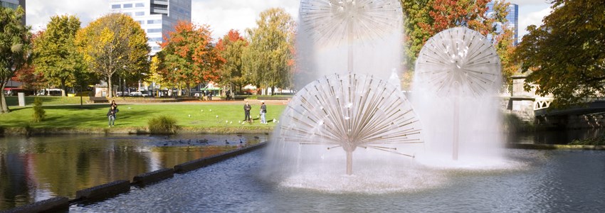A sunny autumn day in Christchurch, New Zealand, with the Ferrier Fountain, the Avon River, tourists chatting and feeding ducks in background, and trees in autumn colours.