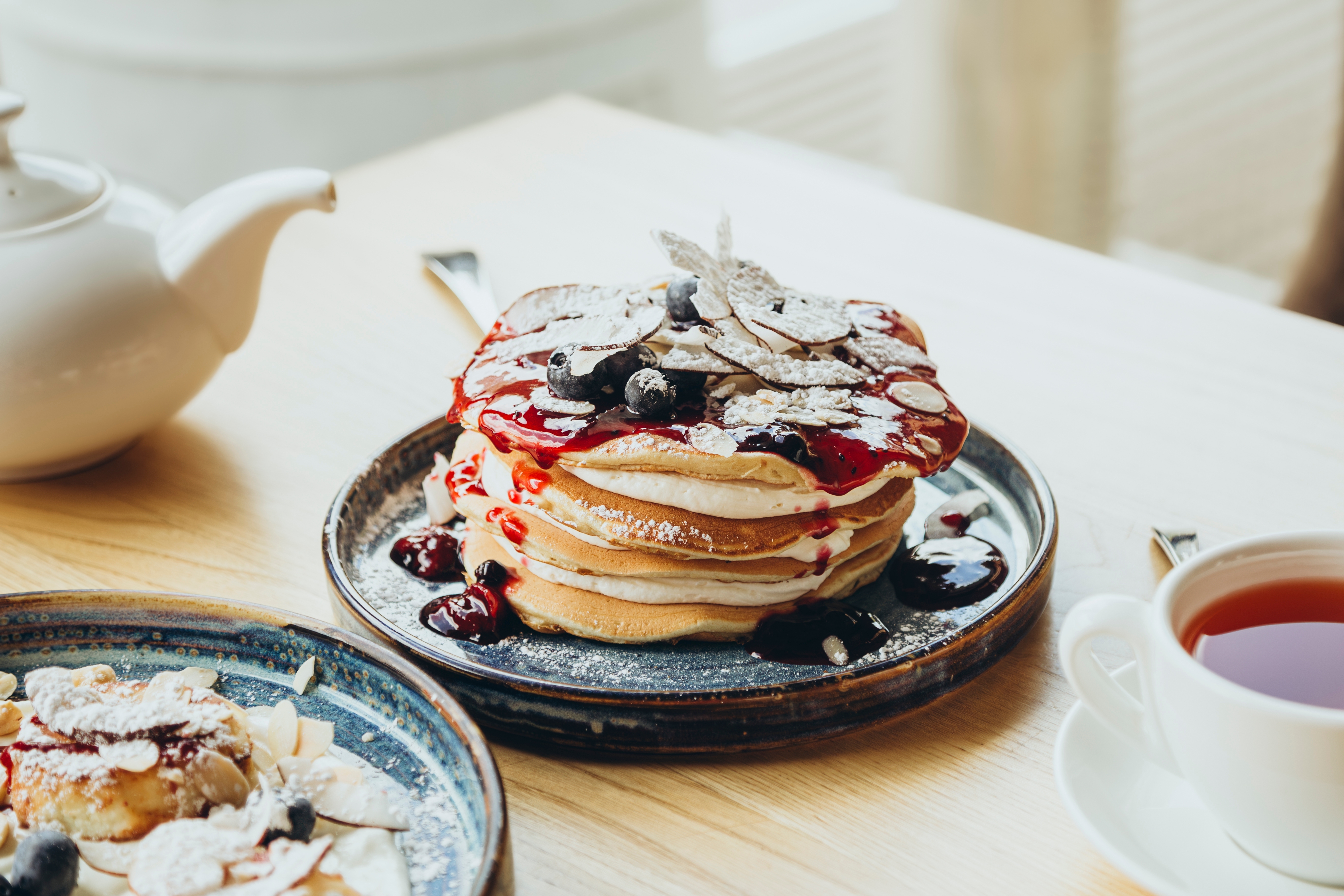 A tall stack of pancakes topped with coconut slices and blueberries, drizzled with thick jam. Breakfast pancakes with berries in cafe. Breakfast served on a wooden table, tea and pancakes.