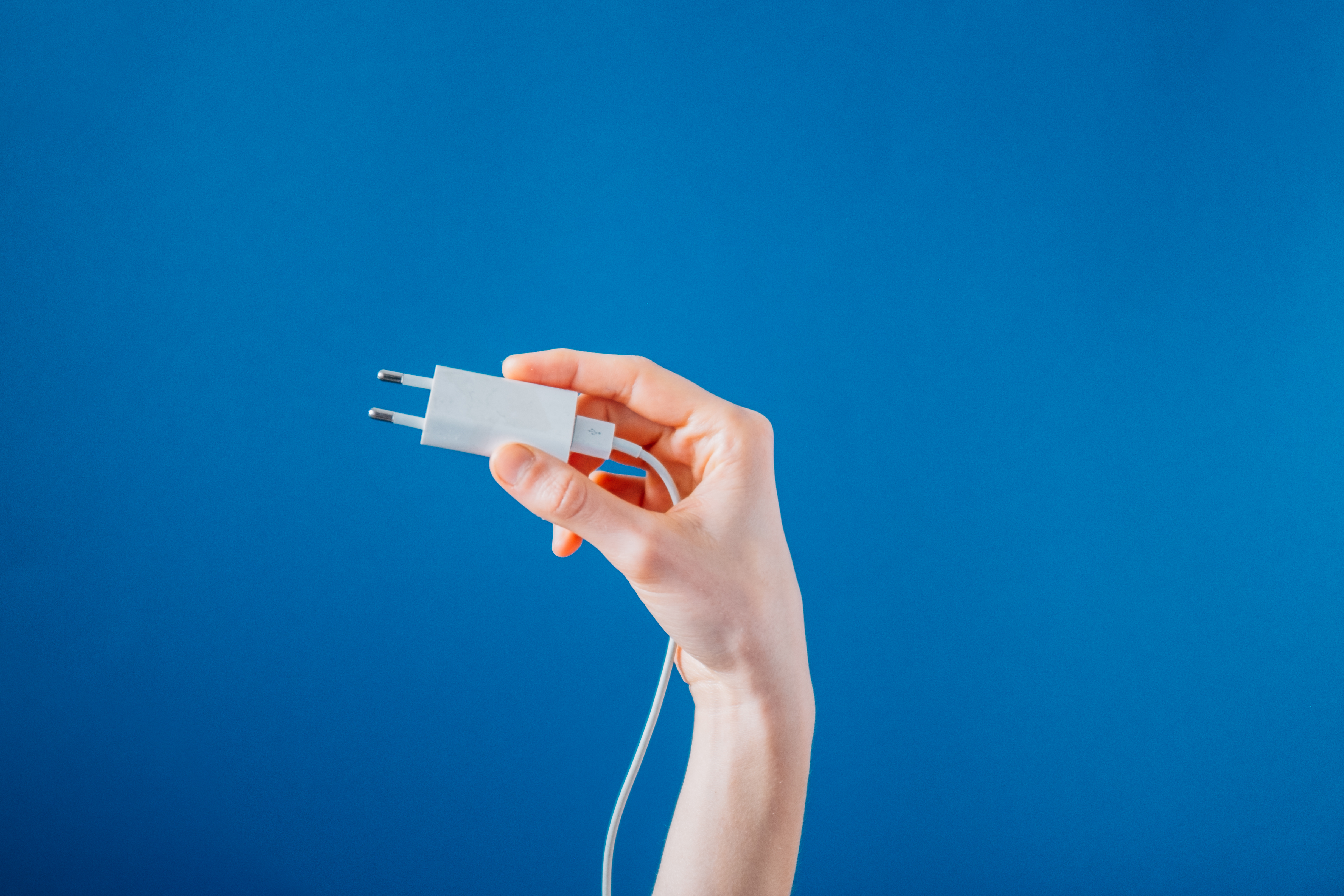 Holding A White Phone Charger On Blue Photo A hand holds up a white European phone charger, the cord dangling off down the arm and the background blue as the skies.