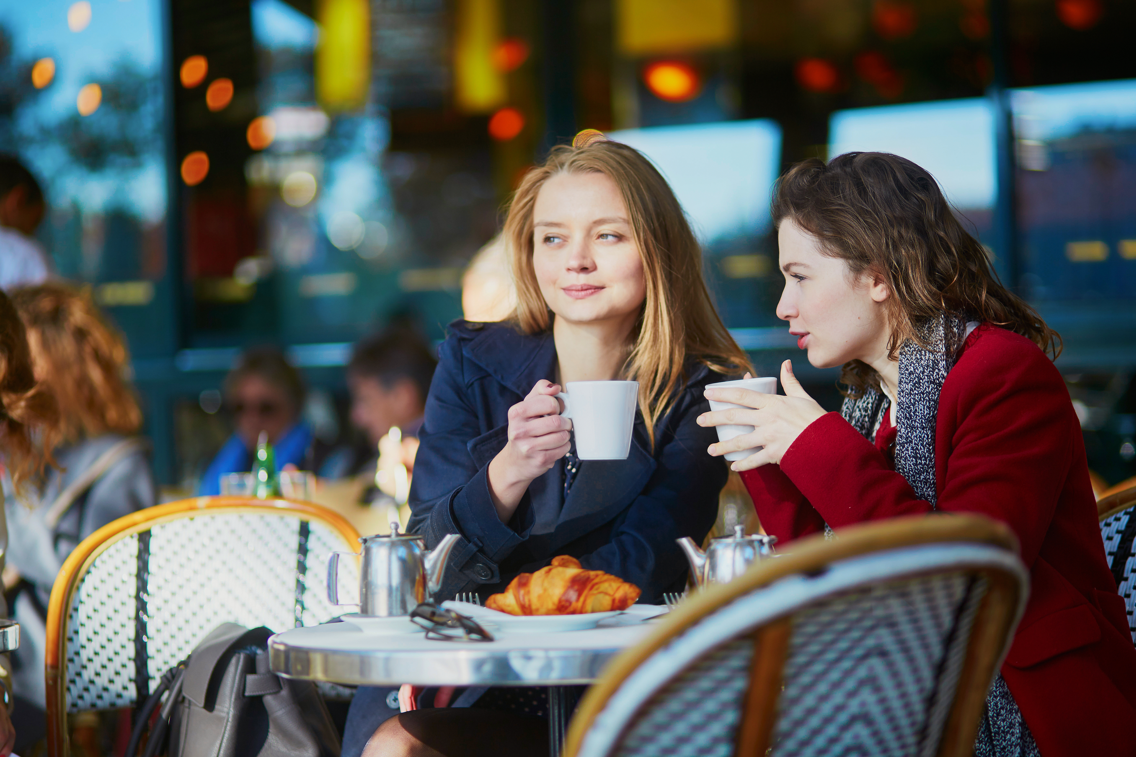 Two young girls in Parisian outdoor cafe, drinking coffee with croissant and chatting. Friendship concept