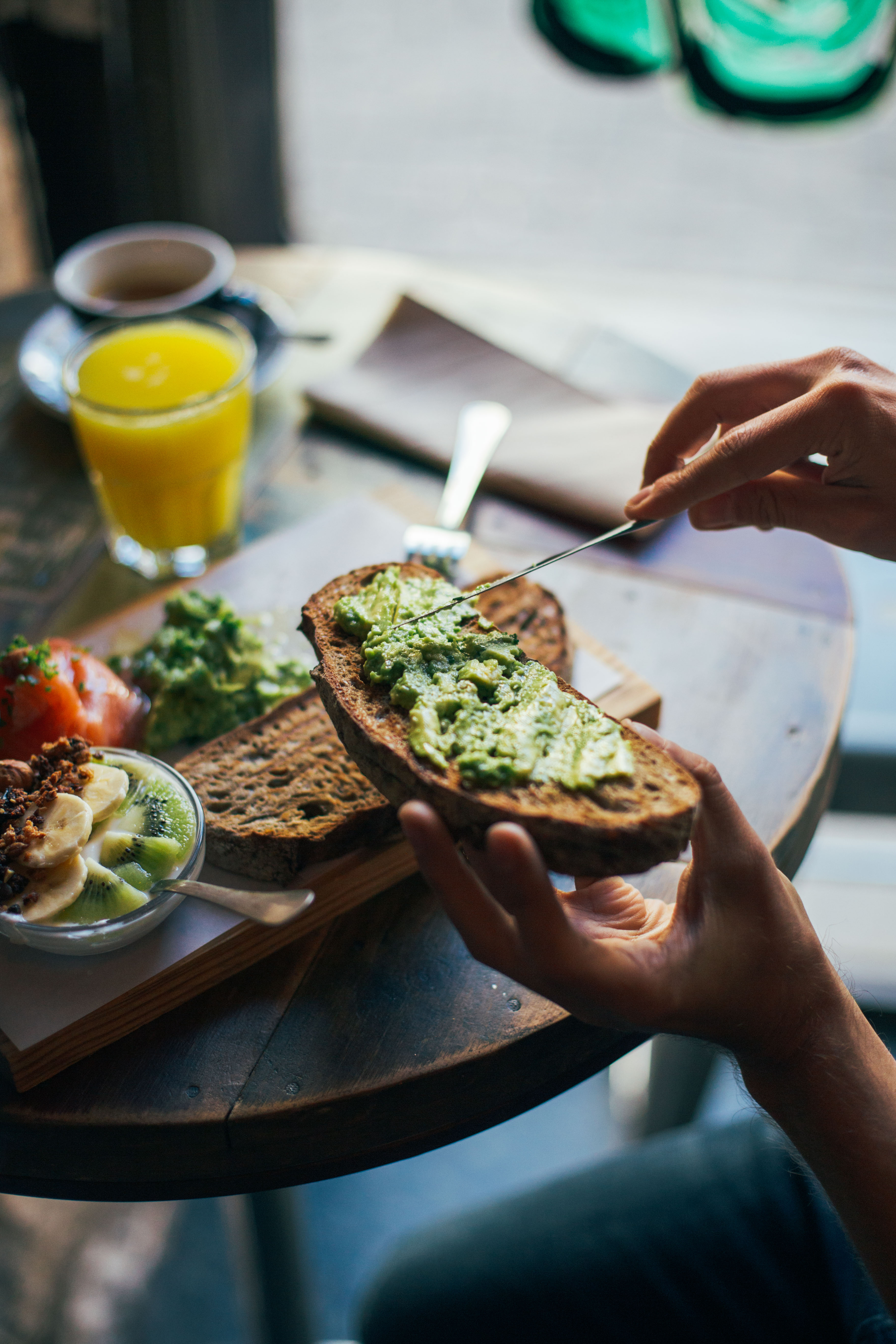 Man spreads avocado on crunchy toasted loaf of bread, breakfast at restaurant or cool coffee spot, with fresh orange juice and other spreads or toppings in background. Enjoy healthy lifestyle choice