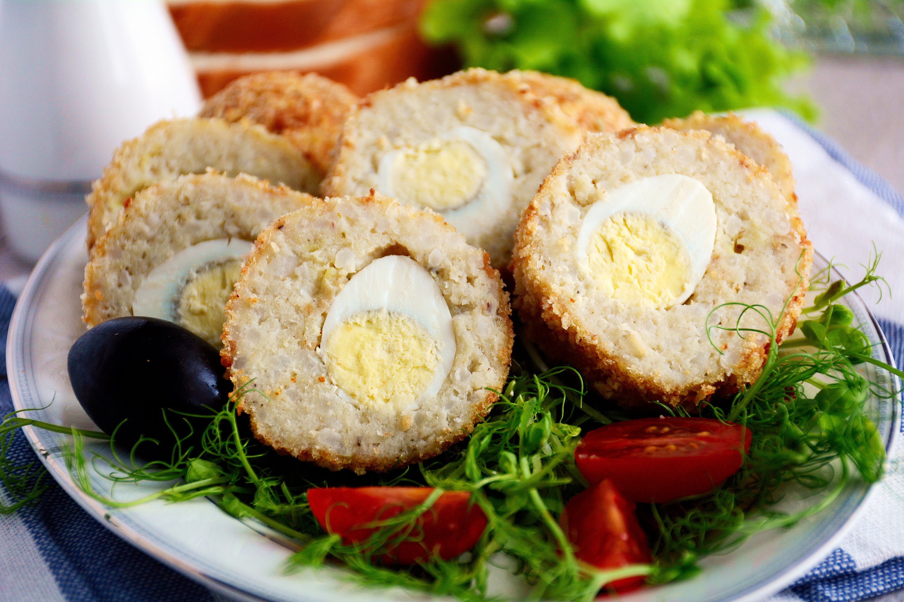 Scottish eggs on white plate with green salad, black olives and cherry tomatoes. Close up