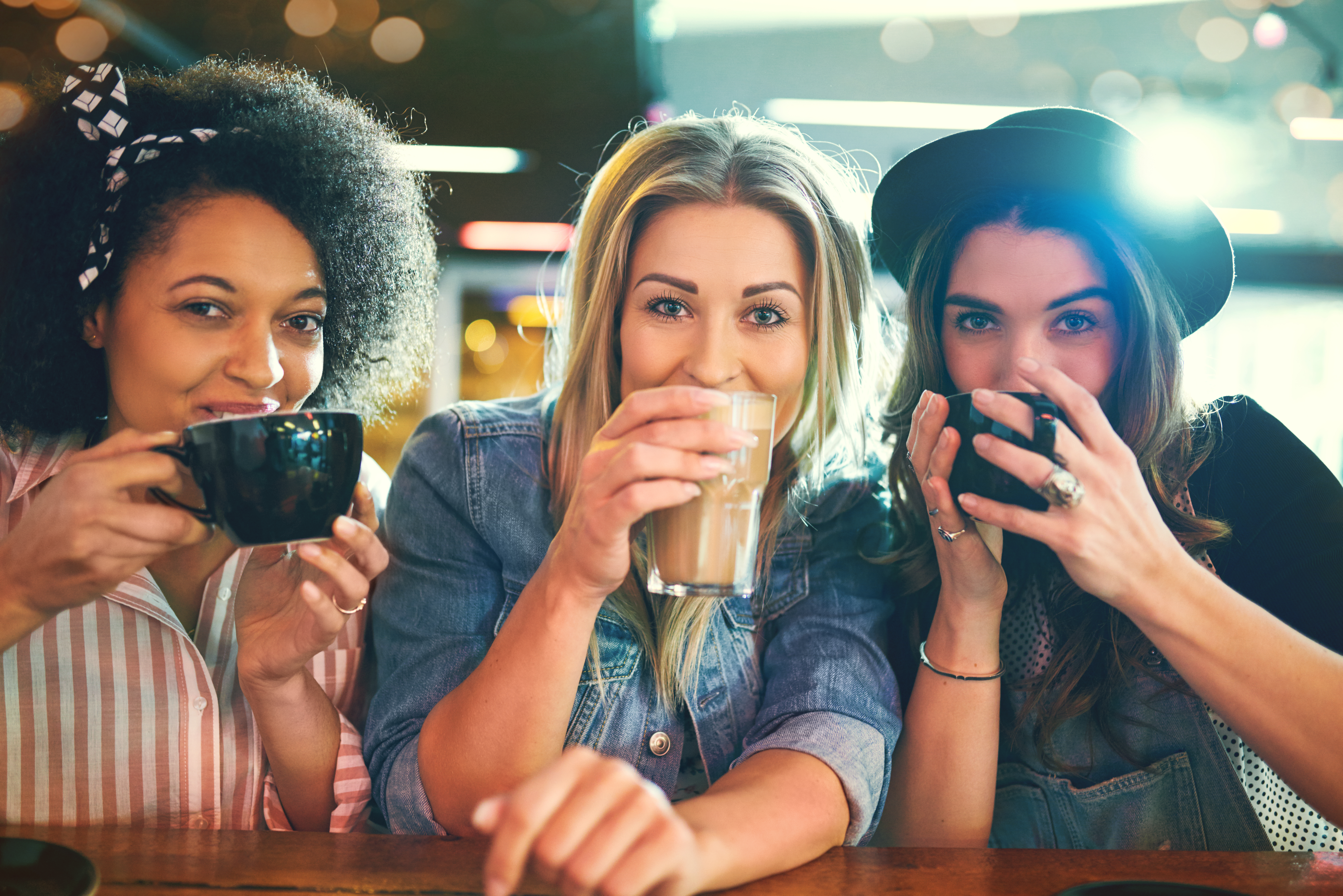 Three young women front portrait close-up at cafe table sipping coffee from their cups, looking at camera and smiling