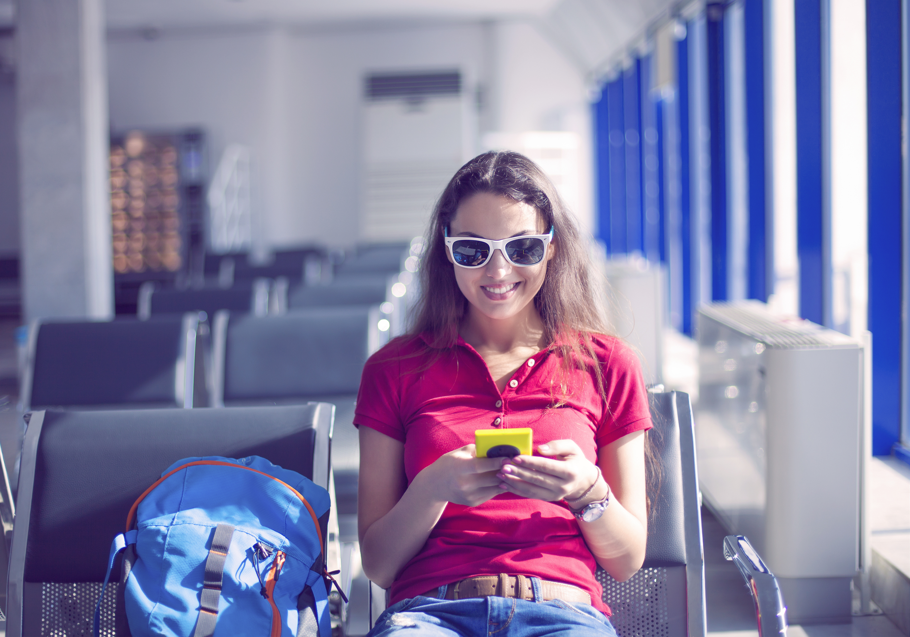 Young female passenger on smart phone at gate waiting in terminal while waiting for her flight. Air travel concept with young casual woman sitting with hand luggage backpack