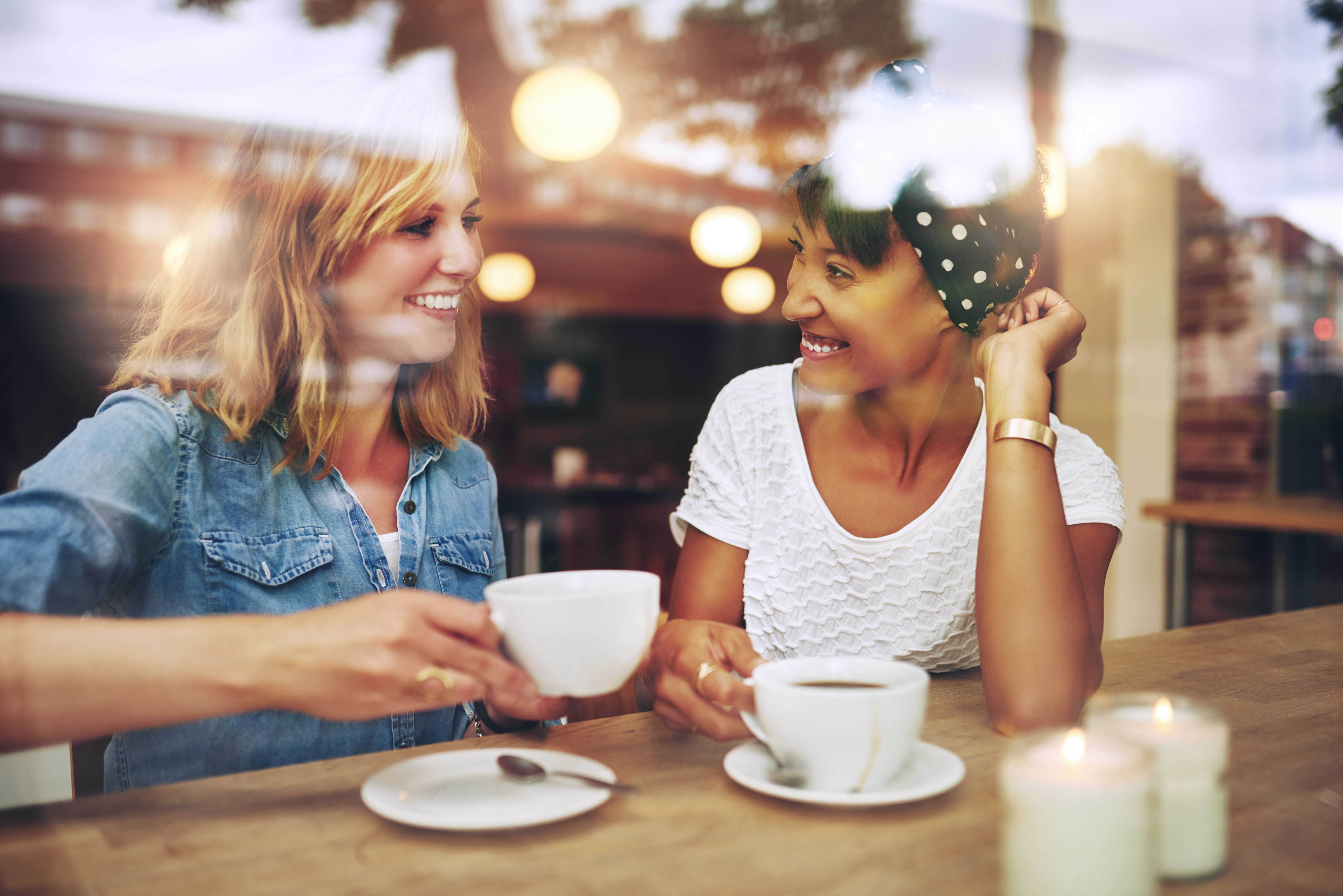 Two multi ethnic friends enjoying coffee together in a coffee shop viewed through glass with reflections as they sit at a table chatting and laughing