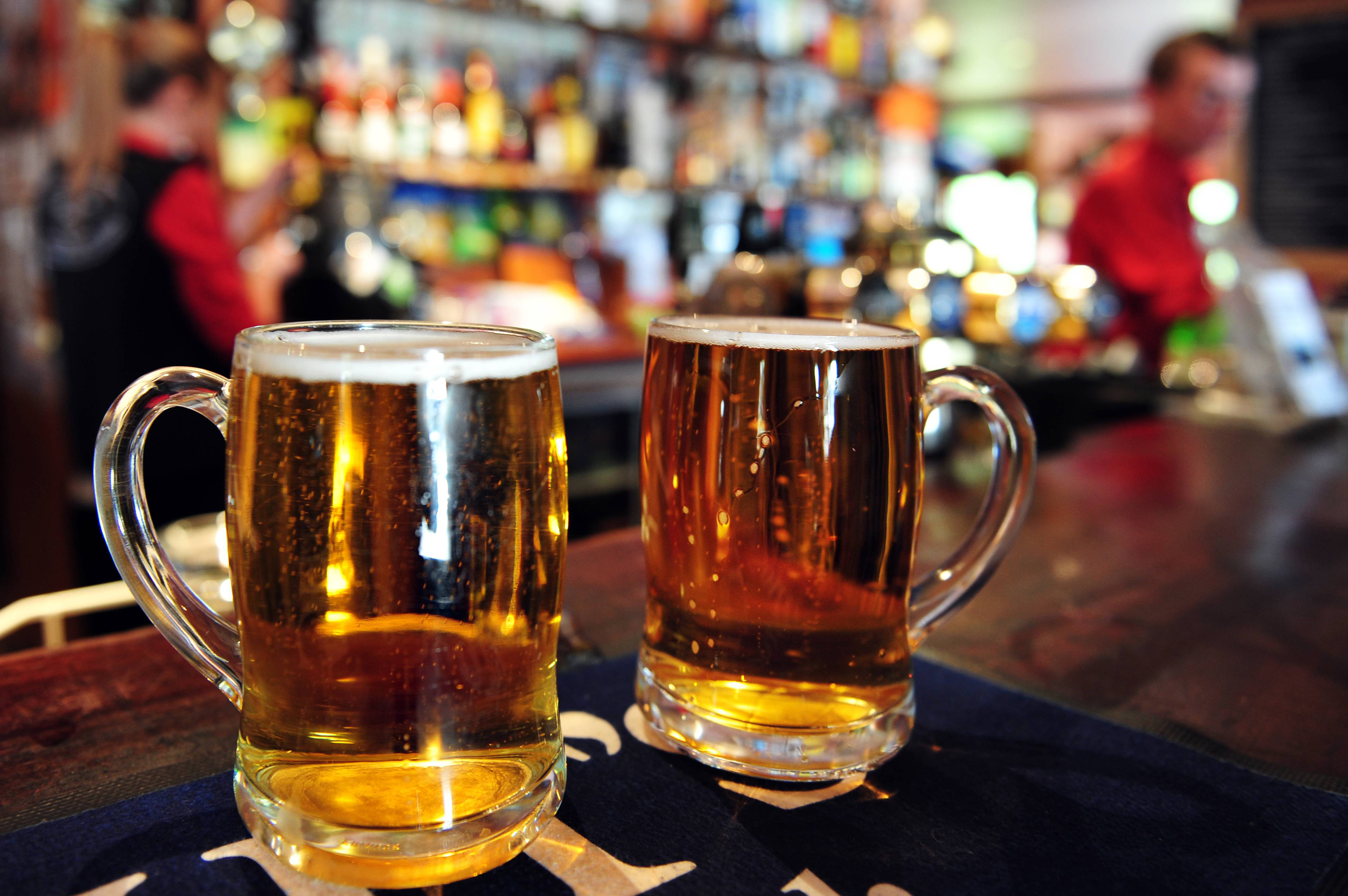 Selective focus close up of two large mugs of cold beer on a pub counter with unrecognizable bar tender in the background. Concept photo of drinking beer and alcohol. Copy space