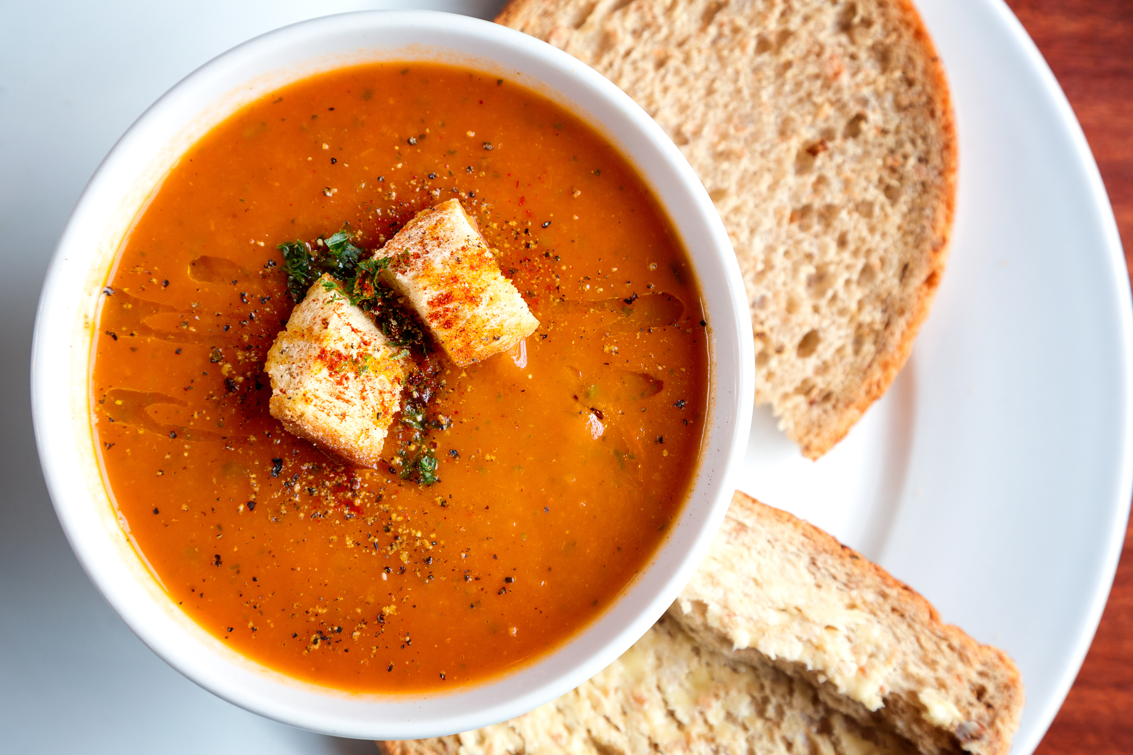 A bowl of fresh tomato soup in white ceramic bowl, garnished with herbs, croutons, seasoning and a drizzle of olive oil, and served with crusty wholemeal bread.