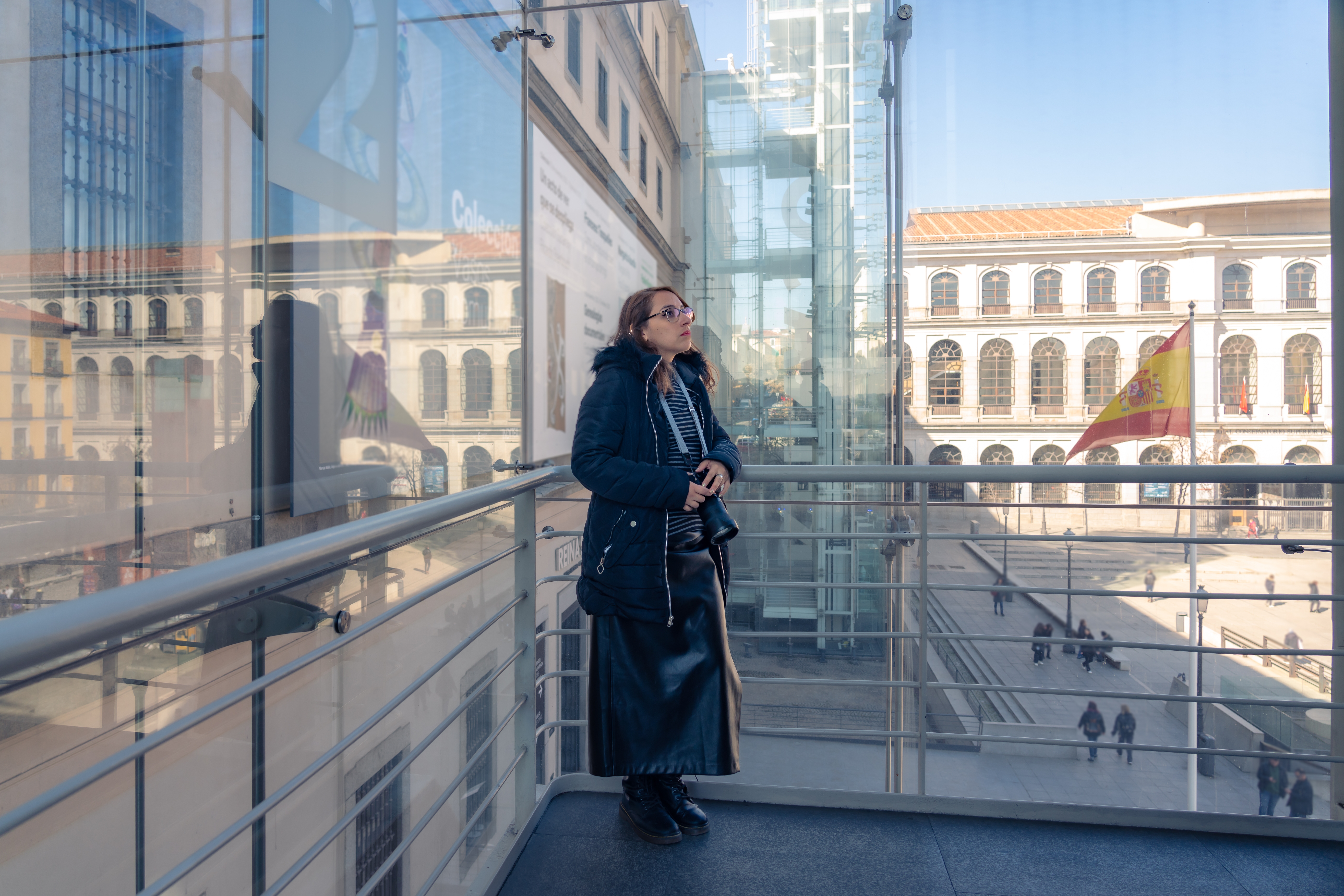 Young Latin woman tourist in warn clothes near window and looking out with professional photo camera in Reina Sofia museum in Madrid, Spain during daytime