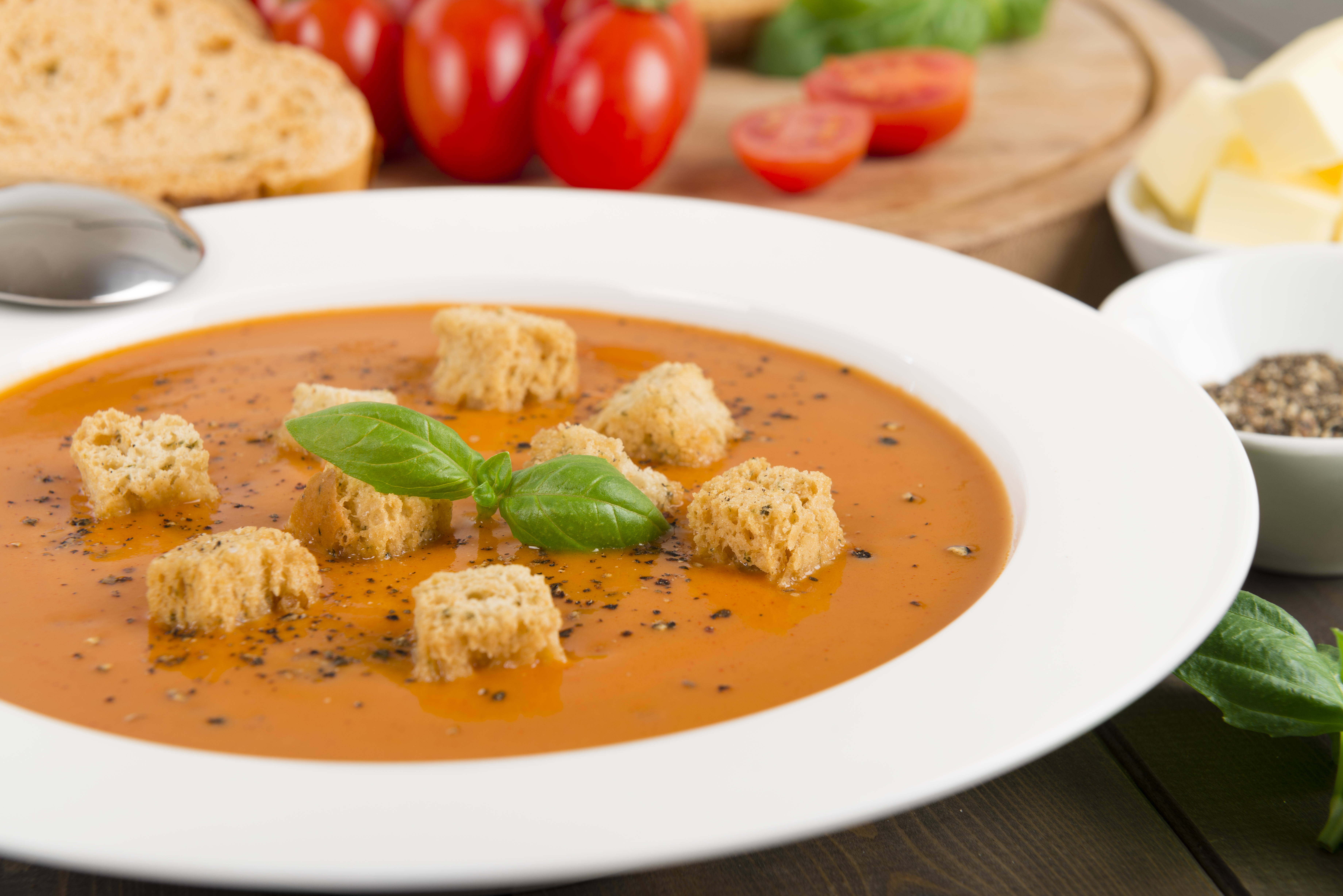 Cream of tomato soup with croutons and cracked black pepper garnished with basil leaves. Sliced crusty bread and butter on background.