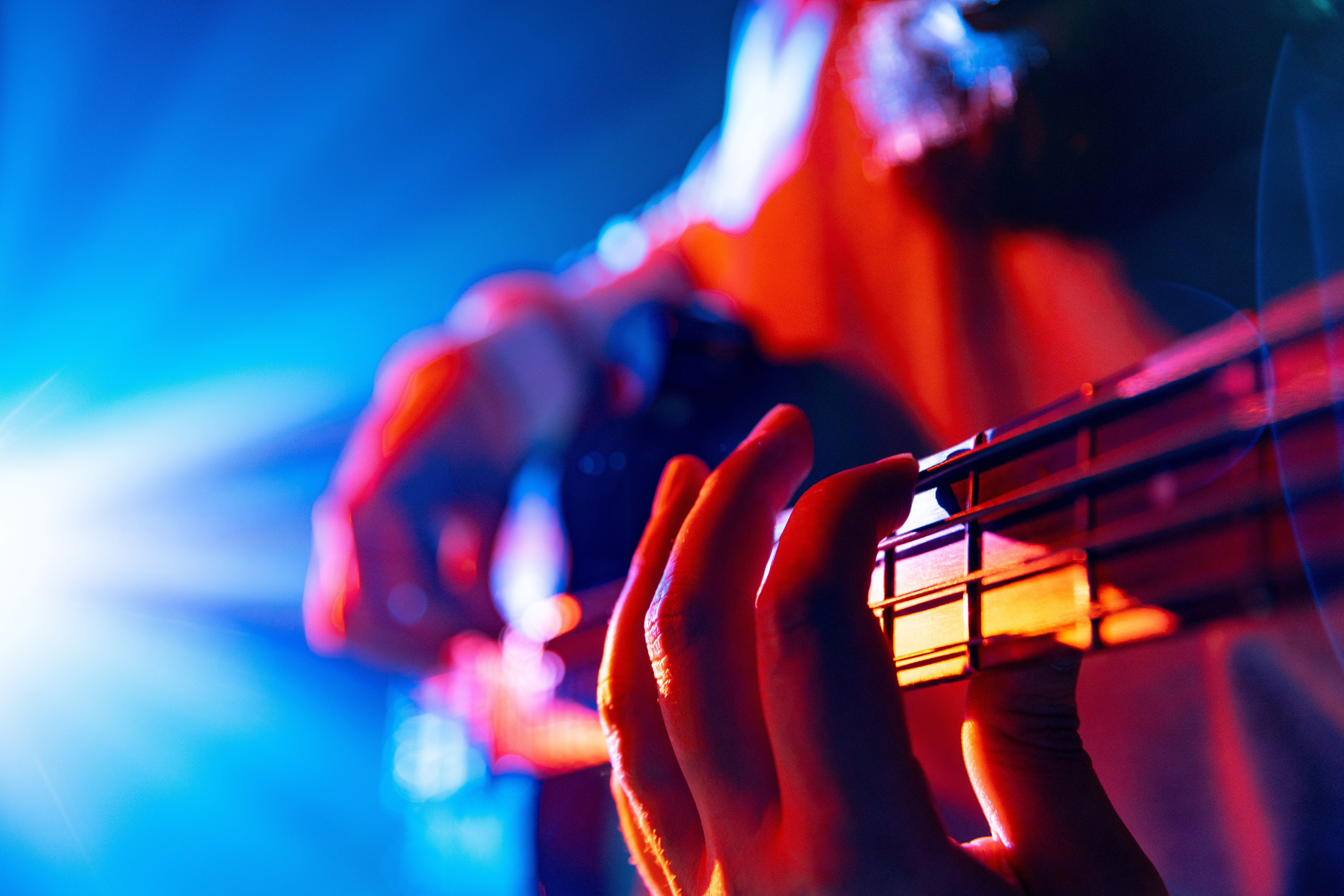 Bassist's hands playing melodic chords of new track in action with colorful concert light flare in background.